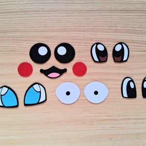 May include: A collection of felt facial features, including black and white eyes, red cheeks, a black mouth with a pink interior, and blue and white eyes. These craft supplies are arranged on a light brown wooden surface.