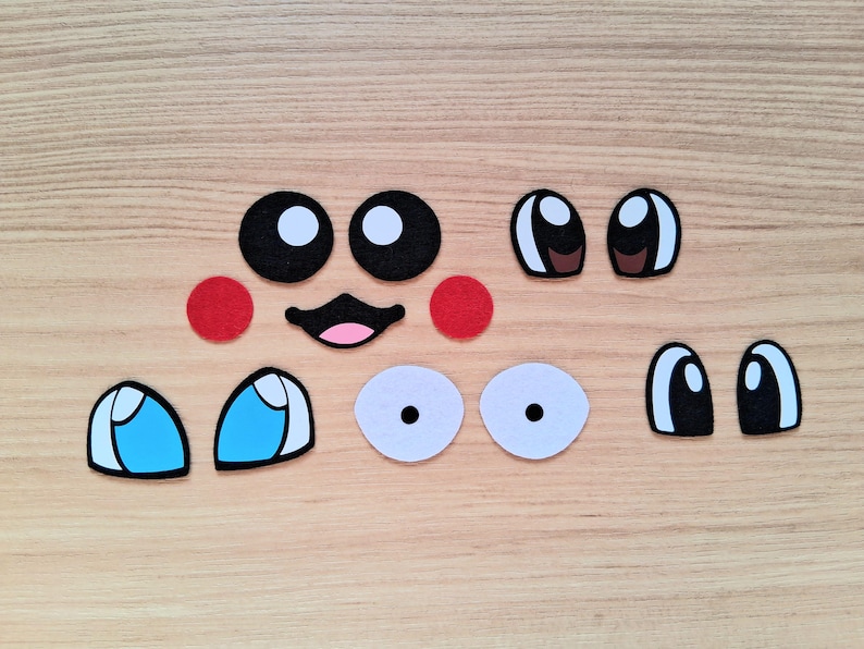 May include: A collection of felt facial features, including black and white eyes, red cheeks, a black mouth with a pink interior, and blue and white eyes. These craft supplies are arranged on a light brown wooden surface.