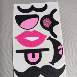 May include: A sheet of pink and black felt stickers with various facial features, including moustaches, lips, eyes, and a tongue.