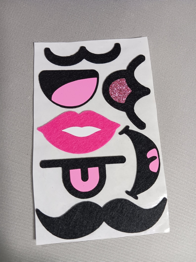 May include: A sheet of pink and black felt stickers with various facial features, including moustaches, lips, eyes, and a tongue.
