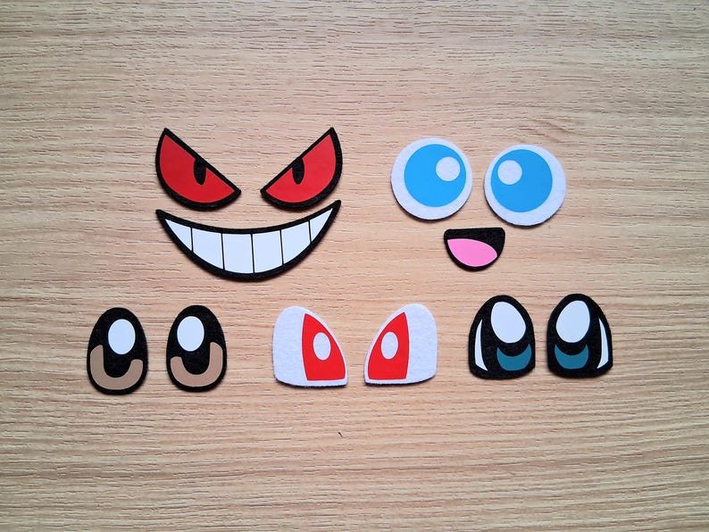 May include: A collection of colorful, cartoon-style face elements, including red and white eyes, a white-toothed grin, blue-eyed circles, and a pink mouth. These craft supplies are arranged on a light brown wooden surface.