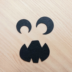 May include: Black paper cutouts of eyes and a mouth on a light wood background, creating a spooky face.