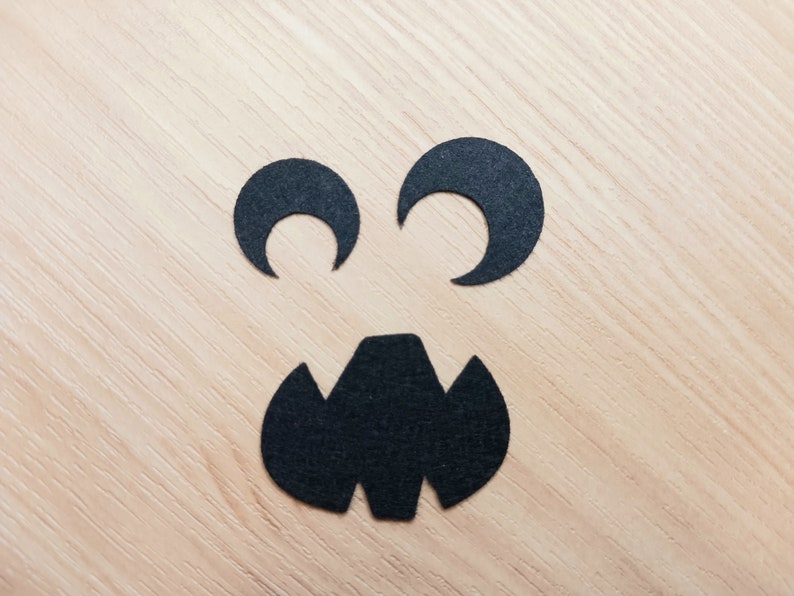 May include: Black paper cutouts of eyes and a mouth on a light wood background, creating a spooky face.