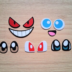 May include: Assortment of cartoon face cutouts, including red and black angry eyes, a smiling mouth, blue eyes, a pink mouth, and various eye shapes. The cutouts are arranged on a light brown wooden surface.