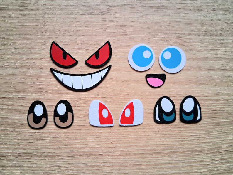 May include: Assortment of cartoon face cutouts, including red and black angry eyes, a smiling mouth, blue eyes, a pink mouth, and various eye shapes. The cutouts are arranged on a light brown wooden surface.