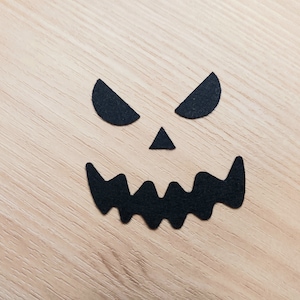 May include: Black paper cutouts of a scary face on a light wood background. The face has two eyes, a nose, and a wide, jagged mouth.