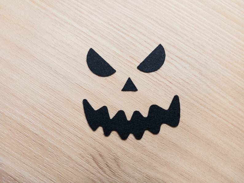 May include: Black paper cutouts of a scary face on a light wood background. The face has two eyes, a nose, and a wide, jagged mouth.