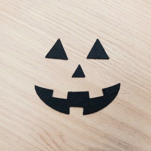 May include: Black paper cutouts of a jack-o'-lantern face on a wood background.
