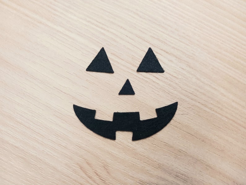 May include: Black paper cutouts of a jack-o'-lantern face on a wood background.