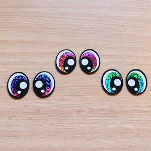 May include: Assortment of colorful googly eyes with iridescent glitter. The eyes are in various colors, including pink, purple, blue, and green.