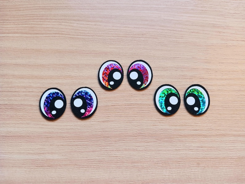 May include: Assortment of colorful googly eyes with iridescent glitter. The eyes are in various colors, including pink, purple, blue, and green.