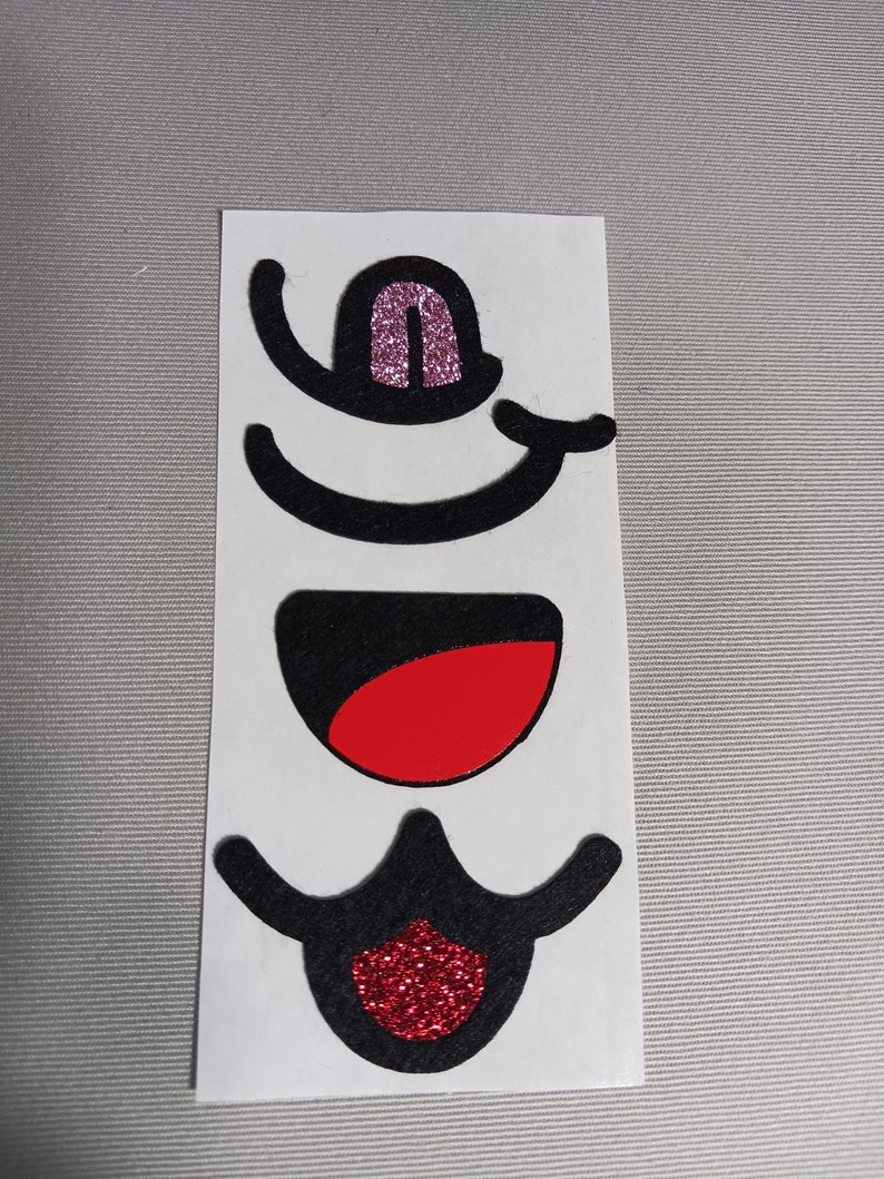 May include: A set of three black felt face stickers with glitter accents. The stickers include a mouth, a nose, and a pair of eyes. The mouth is red, the nose is pink, and the eyes are black.