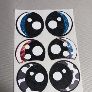 May include: Six googly eye stickers on a white sheet of paper. The eyes are black with white pupils and different coloured accents. The accents include blue glitter, iridescent foil, and a black and white cow print.