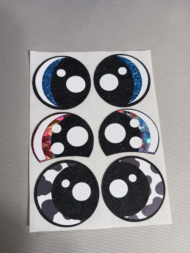 May include: Six googly eye stickers on a white sheet of paper. The eyes are black with white pupils and different coloured accents. The accents include blue glitter, iridescent foil, and a black and white cow print.