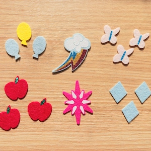 May include: Nine felt appliques in various shapes and colors, including a yellow balloon, a blue cloud with a rainbow, three pink butterflies, three red apples, a pink star, and three blue diamonds.