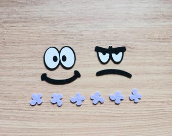 May include: Two felt faces, one smiling with big eyes and the other frowning with furrowed brows. Six small purple felt flowers are arranged below the faces.