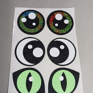 May include: Six colourful felt eye stickers. Two are round with glitter and rainbow colours. Two are black with white circles and a crescent moon shape. Two are green with a black outline and a white circle in the centre.