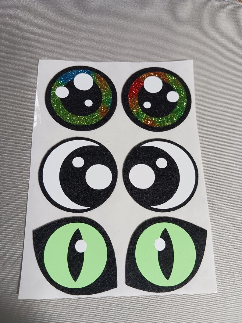 May include: Six colourful felt eye stickers. Two are round with glitter and rainbow colours. Two are black with white circles and a crescent moon shape. Two are green with a black outline and a white circle in the centre.