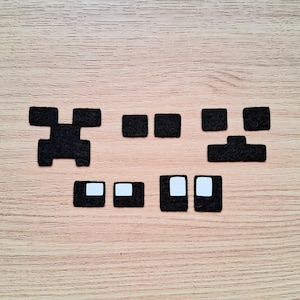 May include: Arrangement of black felt pieces with white square accents, resembling a pixelated character face. The pieces are laid out on a light brown wooden surface, suggesting a craft or DIY project.