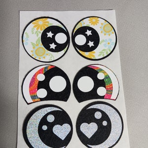 May include: Six colourful eye patches with different designs. The designs include floral, rainbow, and glitter moon patterns. Each patch has a white circle in the centre.