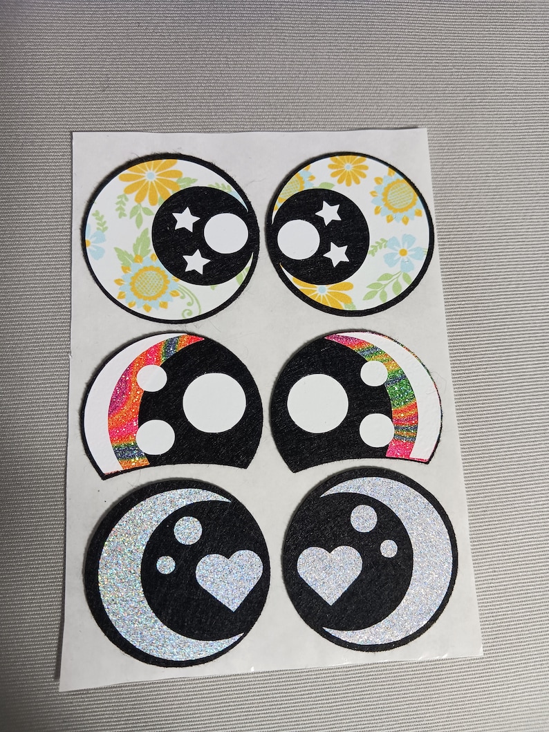 May include: Six colourful eye patches with different designs. The designs include floral, rainbow, and glitter moon patterns. Each patch has a white circle in the centre.