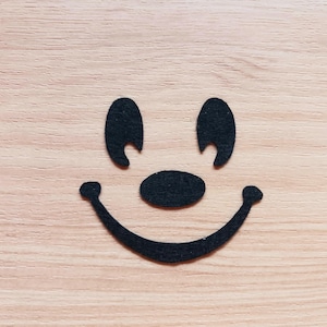May include: A simple, black felt face with two curved eyebrows, an oval nose, and a smiling mouth. The face is set against a light brown wooden background.