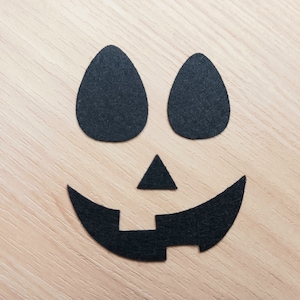 May include: Black felt cutouts of a jack-o'-lantern face on a light wood background. The face has two oval eyes, a triangular nose, and a wide, curved mouth with a small rectangle in the center.