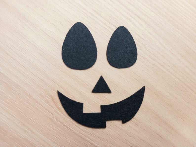 May include: Black felt cutouts of a jack-o'-lantern face on a light wood background. The face has two oval eyes, a triangular nose, and a wide, curved mouth with a small rectangle in the center.
