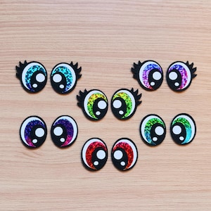 May include: Assortment of colorful googly eyes with black outlines. The eyes are made of glitter and have a variety of colors, including rainbow, green, blue, and purple.