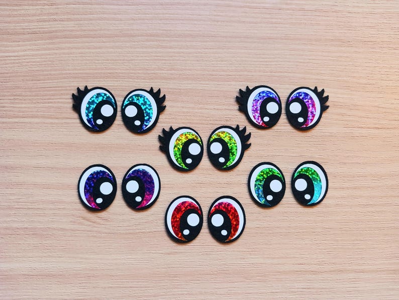 May include: Assortment of colorful googly eyes with black outlines. The eyes are made of glitter and have a variety of colors, including rainbow, green, blue, and purple.
