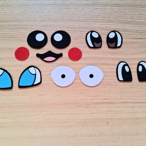 May include: A collection of felt face pieces, including black and white eyes, red circles, a smiling mouth, and blue and white eyes. These craft supplies are arranged on a light wood grain surface.