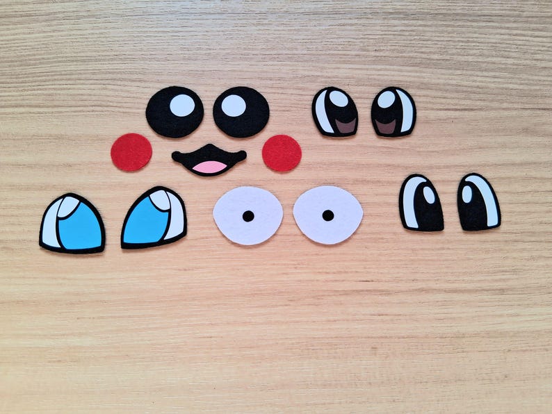 May include: A collection of felt face pieces, including black and white eyes, red circles, a smiling mouth, and blue and white eyes. These craft supplies are arranged on a light wood grain surface.