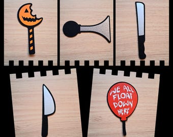 May include: Five Halloween-themed cutouts on wooden blocks. Includes a bitten pumpkin lollipop, a black and silver horn, a silver and black knife, a silver and black knife, and a red balloon with the text "WE ALL FLOAT DOWN HERE".
