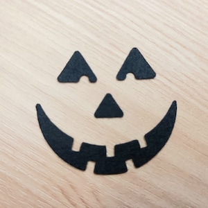 May include: Black paper cutouts of a jack-o'-lantern face on a light wood background.
