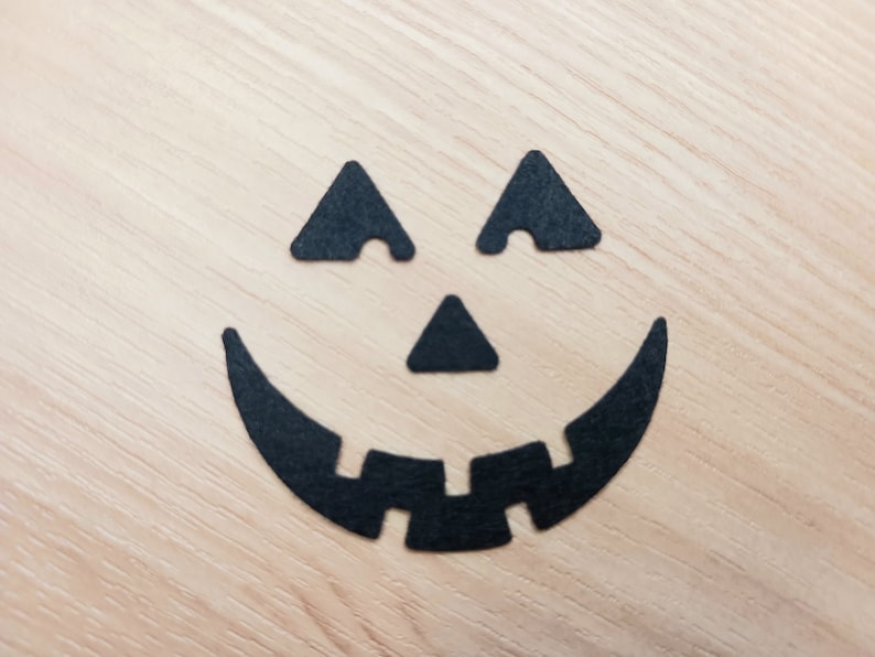 May include: Black paper cutouts of a jack-o'-lantern face on a light wood background.