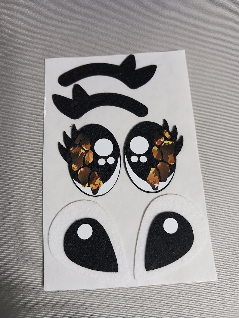 May include: A set of felt eye appliques for crafts. The set includes two pairs of eyes, one pair with a leopard print design and the other pair with a black and white design. The set also includes two pairs of eyelashes.