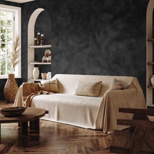 May include: A cozy living room scene featuring a beige sofa draped with a matching throw. Decorative pillows in neutral tones add texture. A wooden coffee table and a woven vase with dried plants complete the warm, inviting space. The walls are dark gray.