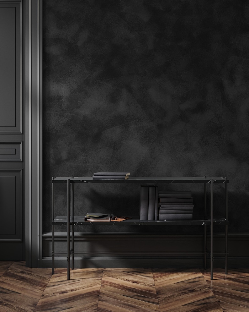 May include: A black metal bookshelf with two shelves, holding stacks of books and a few smaller items. The background features a dark, textured wall and a wooden floor with a herringbone pattern. The room is painted in shades of black and dark brown.