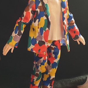 Può includere: Bambola che indossa un completo con stampa floreale colorata, una felpa con cappuccio bianca e scarpe da ginnastica bianche. Il completo presenta un motivo di forme rosse, blu, gialle e marroni. La bambola ha i capelli castani e un'espressione neutra.