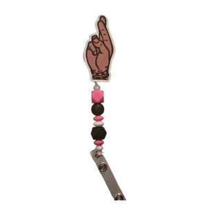 May include: A badge reel with a cartoon hand illustration, fingers crossed. The reel features a pink, black, and white bead design. The hand illustration has a black studded wristband and pink nail polish.