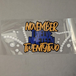 May include: Clear plastic sticker with the words "NOVEMBER TWELVE NINETEEN TWENTY TWO" in orange and blue. A white pearl necklace design is on the sticker.