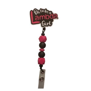 May include: A badge reel with a white and pink design that reads "Oh to be a Lambda Girl." The reel features a black beaded chain with pink and black beads. The badge reel has a silver clip.