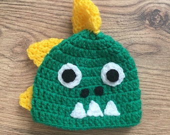 May include: Green crocheted dinosaur hat with yellow spikes, white teeth, and black eyes.