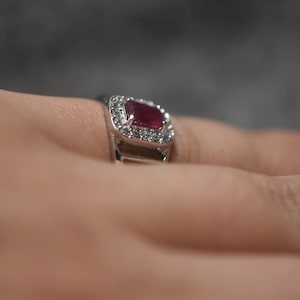 May include: A silver ring featuring a red gemstone surrounded by smaller white gemstones.