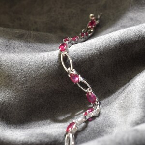 May include: A silver bracelet featuring oval-shaped ruby gemstones set in a delicate design. The bracelet is displayed on a grey fabric background.