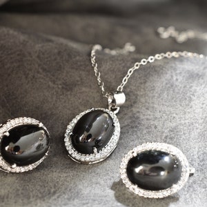 May include: A black onyx jewelry set with a silver chain necklace, earrings, and a ring. Each piece features a black oval stone surrounded by a halo of clear cubic zirconia.