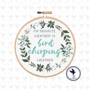 May include: A cross-stitch embroidery hoop with the text "MY FAVORITE WEATHER IS bird chirping WEATHER." The design features a floral wreath in shades of green and pink, with a small logo in the bottom right corner.
