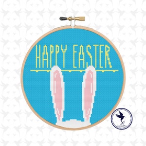Pode incluir: Um aro de bordado azul com o texto "Happy Easter" em letras amarelas. Duas orelhas de coelho cor-de-rosa estão penduradas no aro.