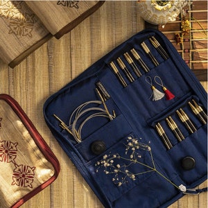May include: A navy blue knitting needle set with gold and black needles, a case, and accessories. The case is open, revealing various knitting tools, including needles, cables, and small decorative tassels. The set is displayed on a wooden surface.