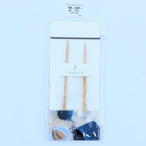 May include: A packet of Seeknit circular knitting needles. The needles are made of light-coloured wood and are size US 6, 100 cm long. The package also contains a small ball of blue yarn, a small plate with biscuits, and a blue fabric with a white design.
